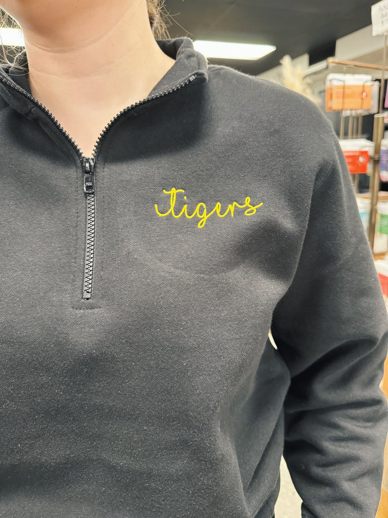 Black fleece pullover with 'Tigers' embroidered in yellow on a blurred indoor background