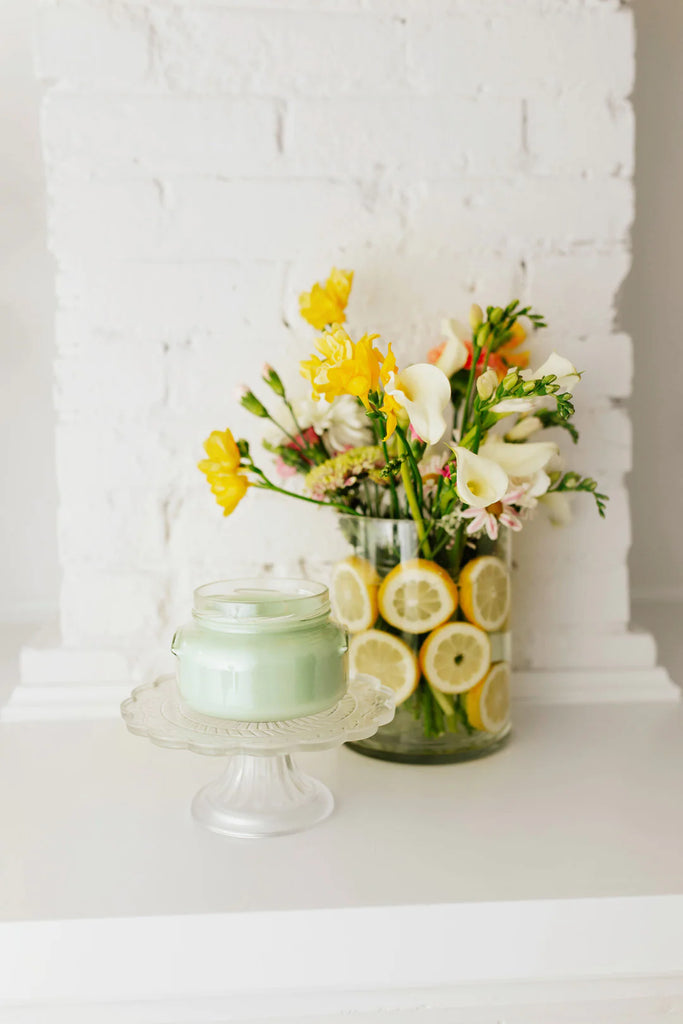 candle on a clear glass stand with a vase of flowers in the back on a white background