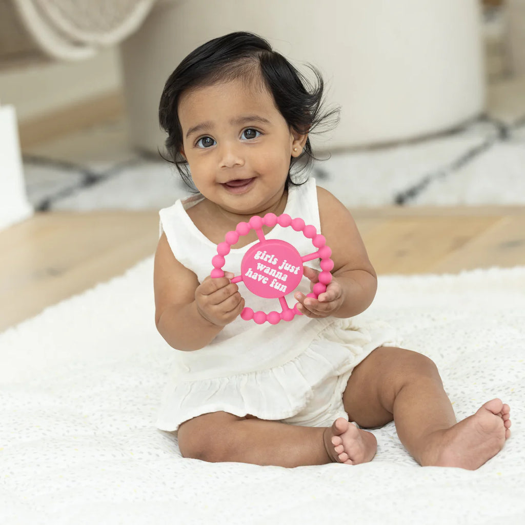 Baby holding a pink teething ring with text, sitting on a white blanket.