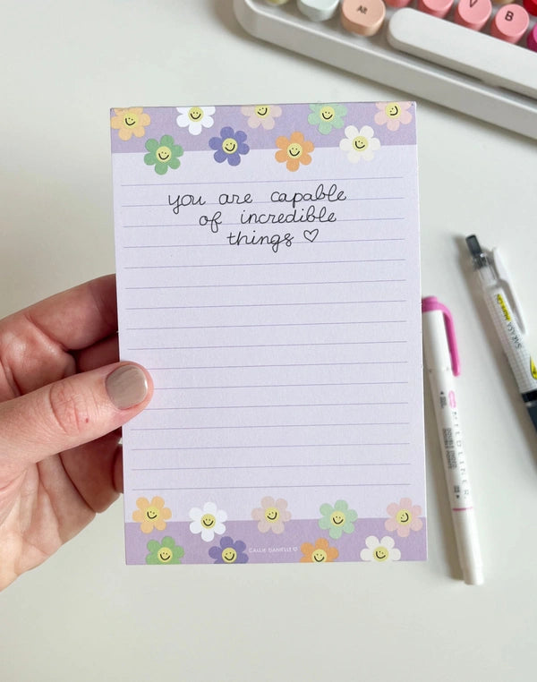 Notepad with motivational quote and floral design held by a hand, with pens and a keyboard in the background.