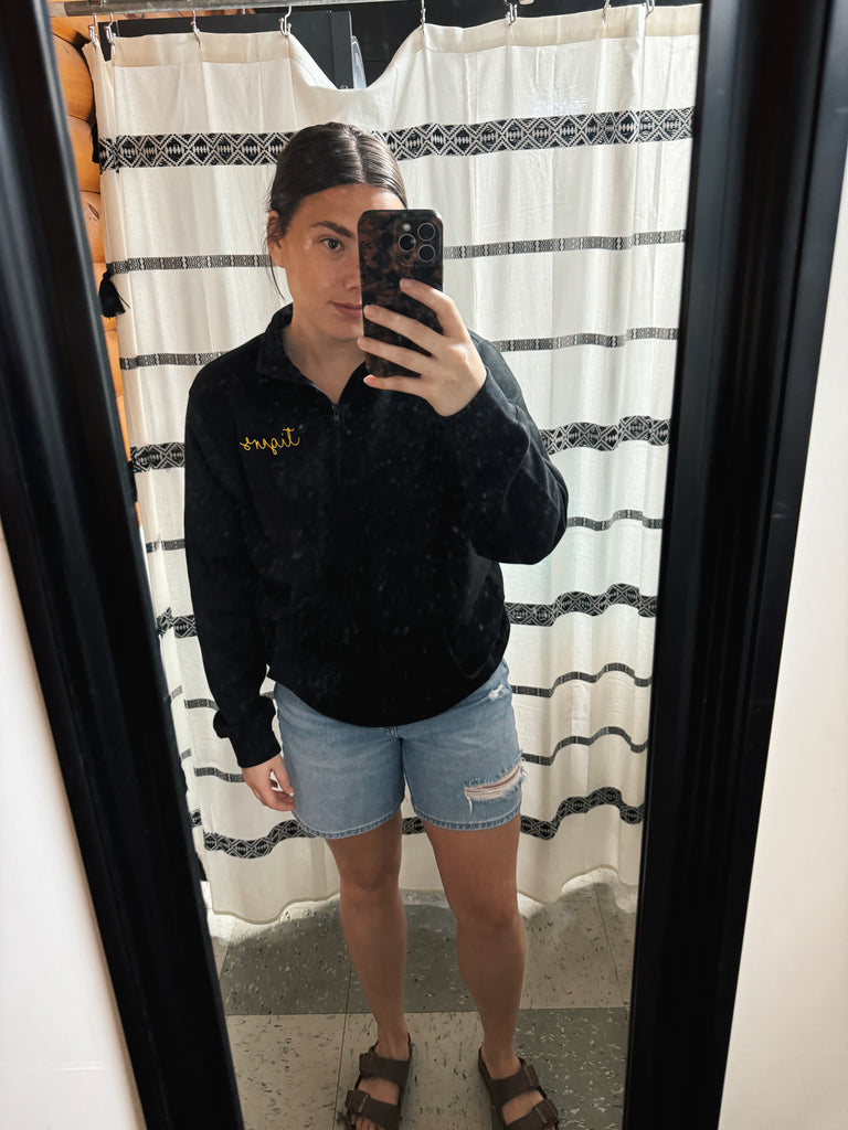 Person taking a mirror selfie wearing a black sweater and denim shorts in front of a striped shower curtain.