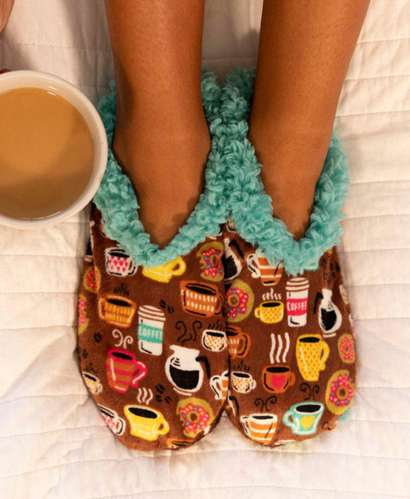 Pair of brown slippers with coffee-themed design and teal trim, worn by a person holding a cup of coffee.