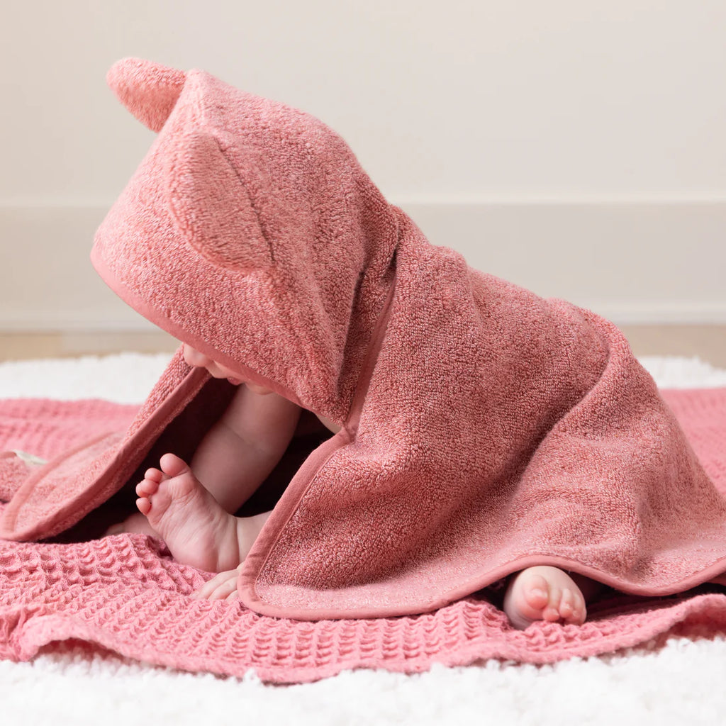 Baby peeking out from under a pink hooded towel on a white surface.