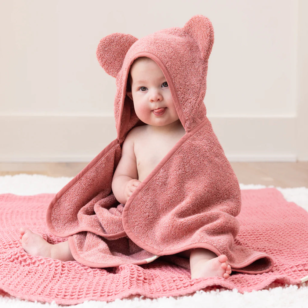 Baby wrapped in a pink hooded towel with bear ears, sitting on a white rug.