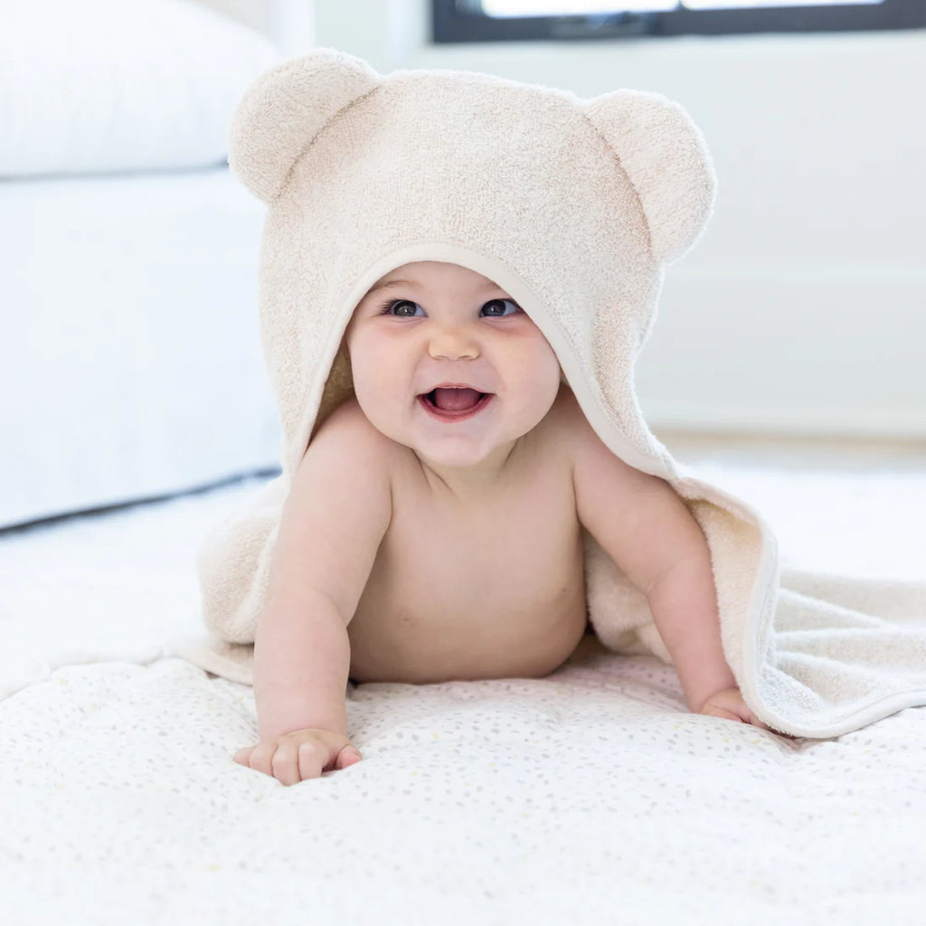Baby wearing a beige hooded towel with bear ears, smiling on a white surface.