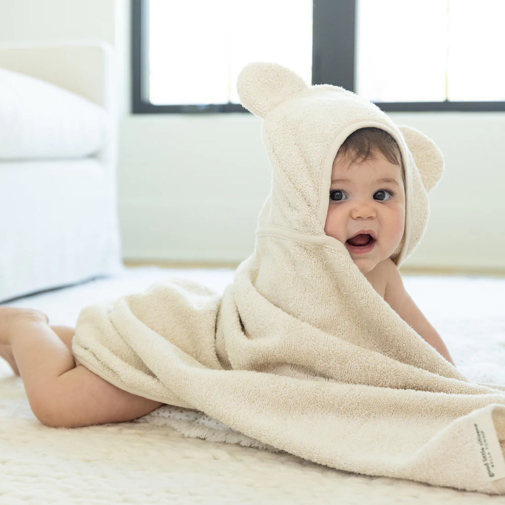 Baby wrapped in a beige towel with bear ears, sitting on a bed.