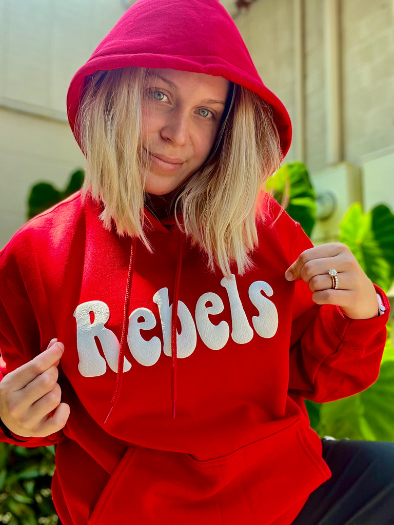 Person wearing a red hoodie with 'Rebels' text, sitting outdoors.