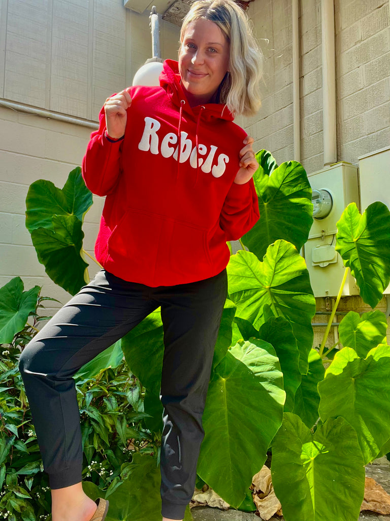 Person wearing a red 'Rebels' hoodie standing outdoors with green plants in the background
