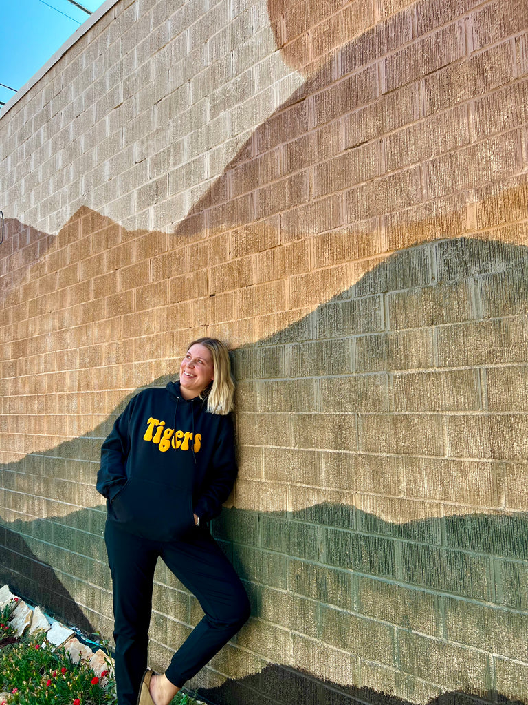 Person wearing a black sweatshirt with 'Tigers' printed on it, leaning against a textured wall.