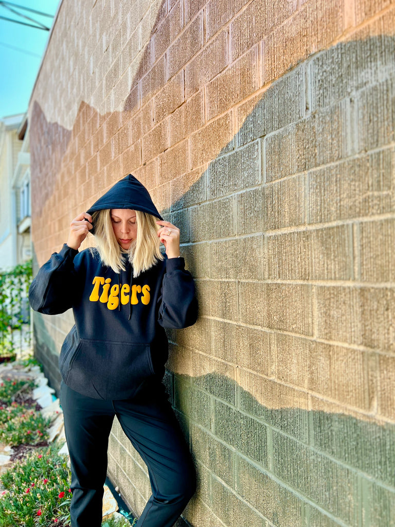 Person wearing a black hoodie with 'Tigers' printed on it, leaning against a brick wall.