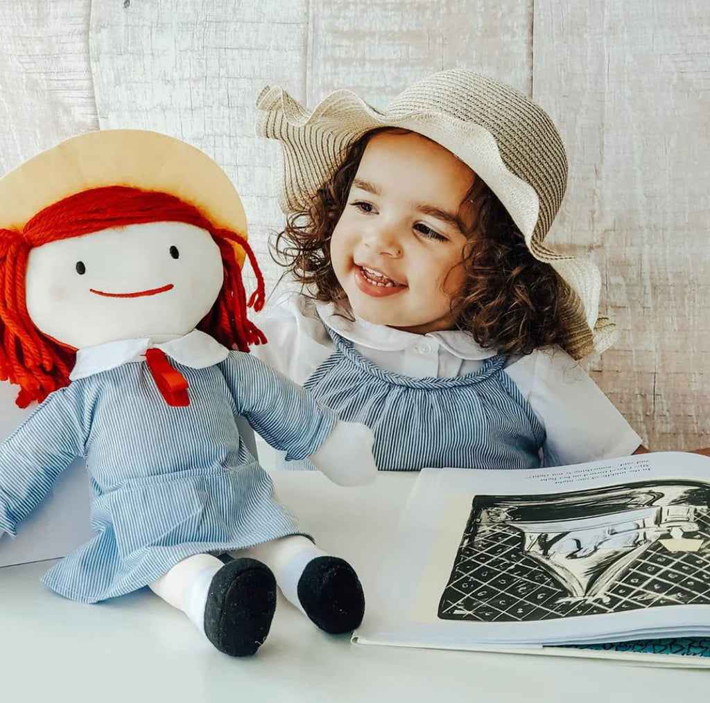 Child in a straw hat with a doll, both wearing blue dresses, sitting at a table with a book.