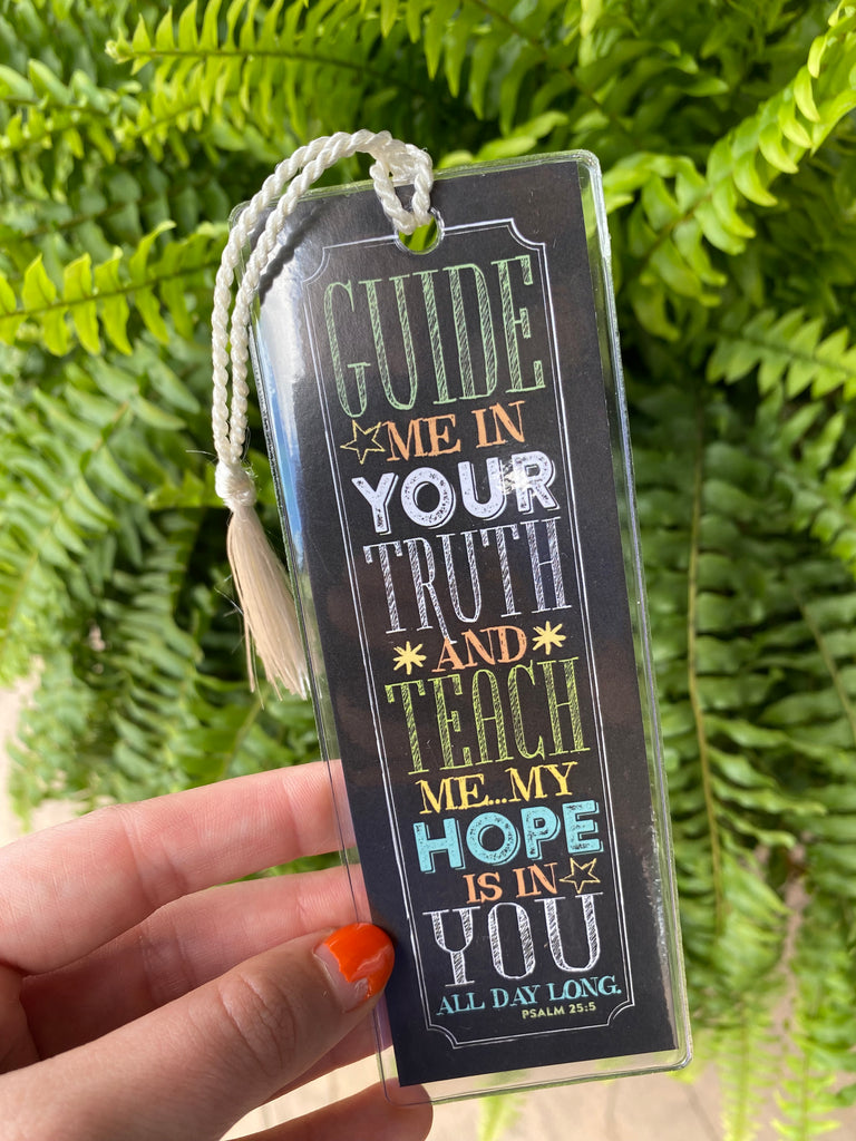 guide me bookmark in a clear plastic covering being held in front of a green fern plant