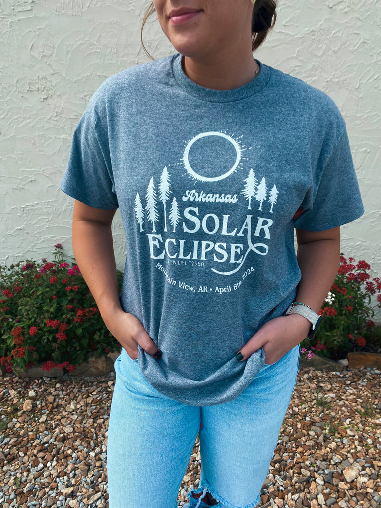 view life 72560's 2024 solar eclipse gray t-shirt with white screen printed arkansas solar eclipse viewlife 72560 mountain view arkansas april 8th, 2024 on a person standing on a river rock ground with small flower bed housing two red flower bushes in front of a white texture painted wall