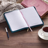 Open notebook with a pen and cup of tea on a wooden surface, with a Bible in the background.