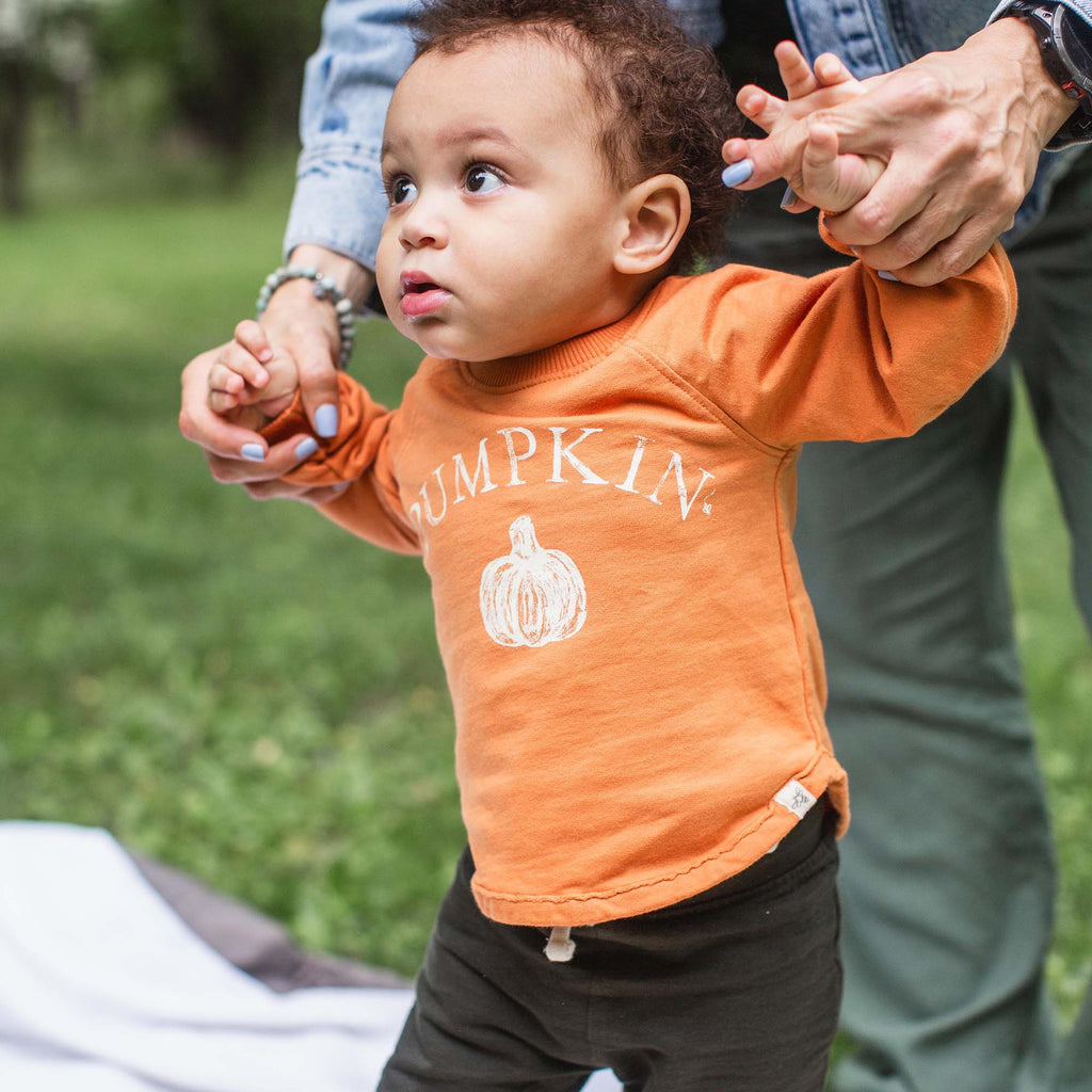 Child wearing an orange shirt with a pumpkin graphic and text, standing outdoors.