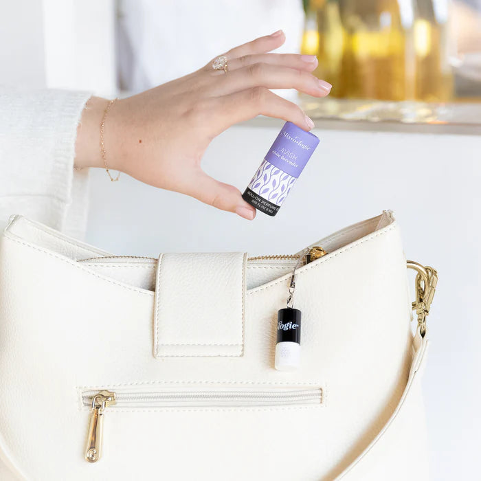 Hand holding a purple rollerball bottle labeled 'Lavender' above a white handbag.