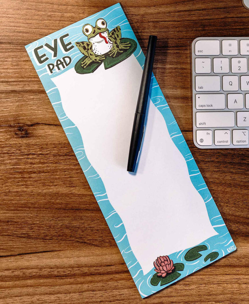 Notepad with a frog design on a wooden desk with a pen and keyboard.