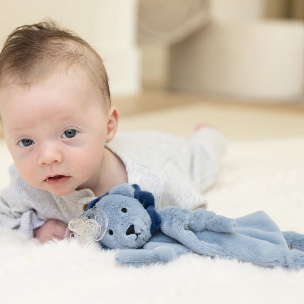 Baby lying on a white blanket with a blue teddy bear and pacifier