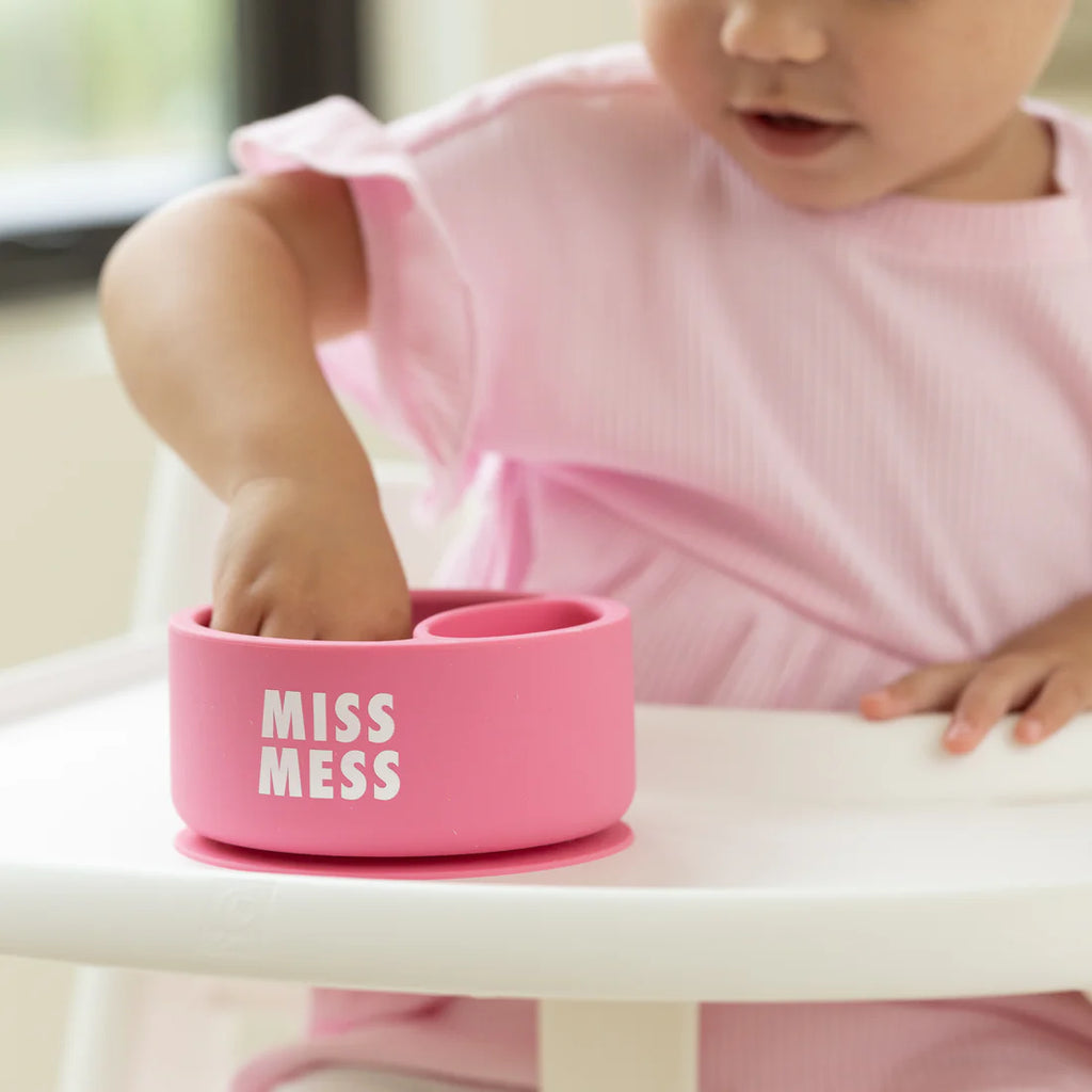 Pink 'MISS MESS' container on a high chair with a child in a pink shirt.