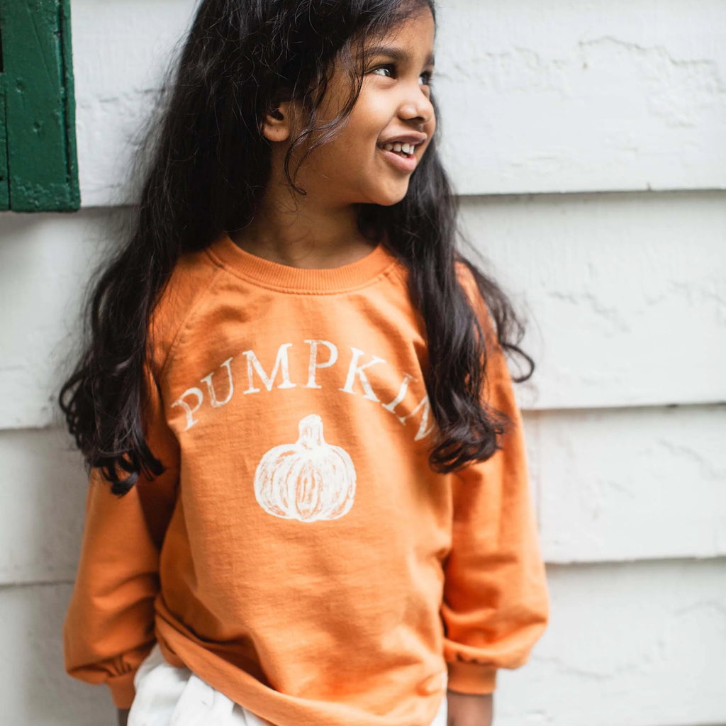 Young girl wearing an orange sweatshirt with 'PUMPKIN' and a pumpkin graphic, standing against a white wall.