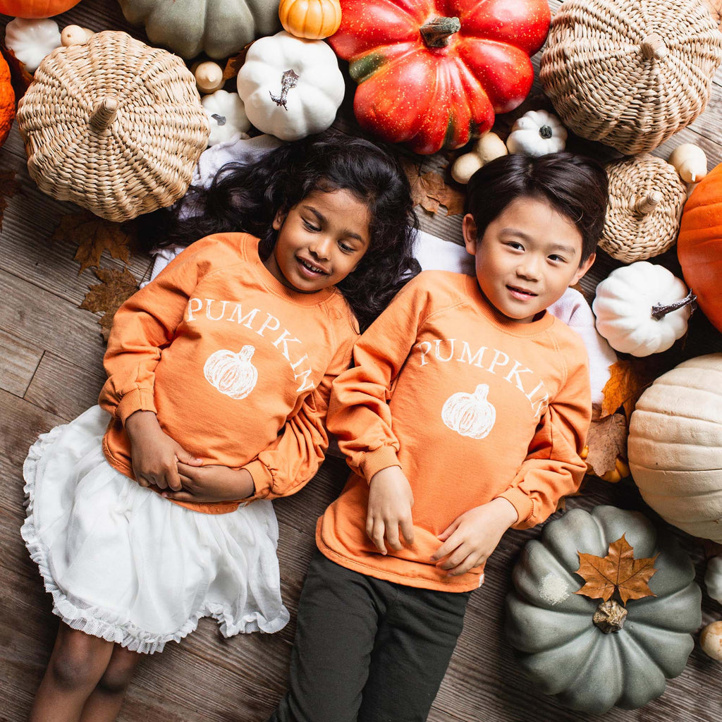 Two children wearing orange 'Pumpkin' shirts surrounded by pumpkins and fall decorations.