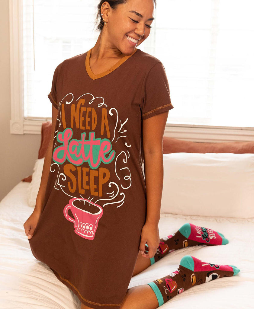 Woman wearing a brown nightshirt with 'I Need a Caffiene Sleep' text and colorful socks on a bed.