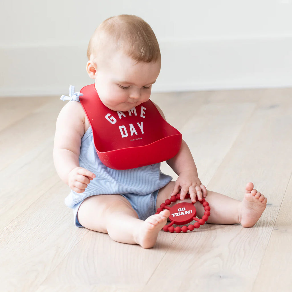 Baby wearing a red bib with 'Game Day' text, sitting on a light wooden floor.