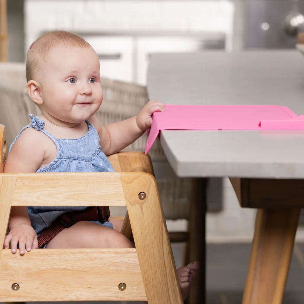 Baby in a wooden high chair with a pink tray, sitting at a table.