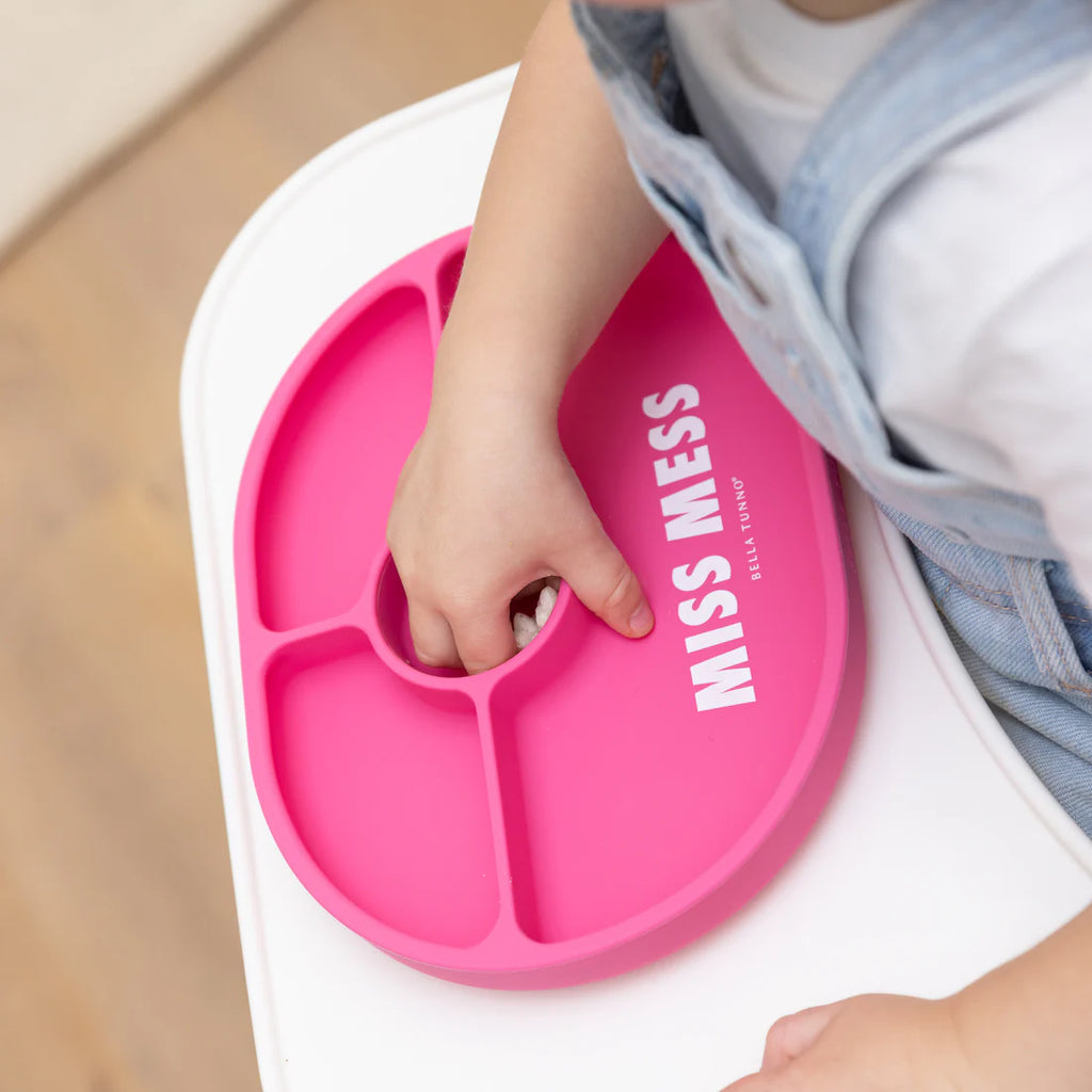 Pink 'Miss Mess' tray with a child's hand reaching for it on a white surface.