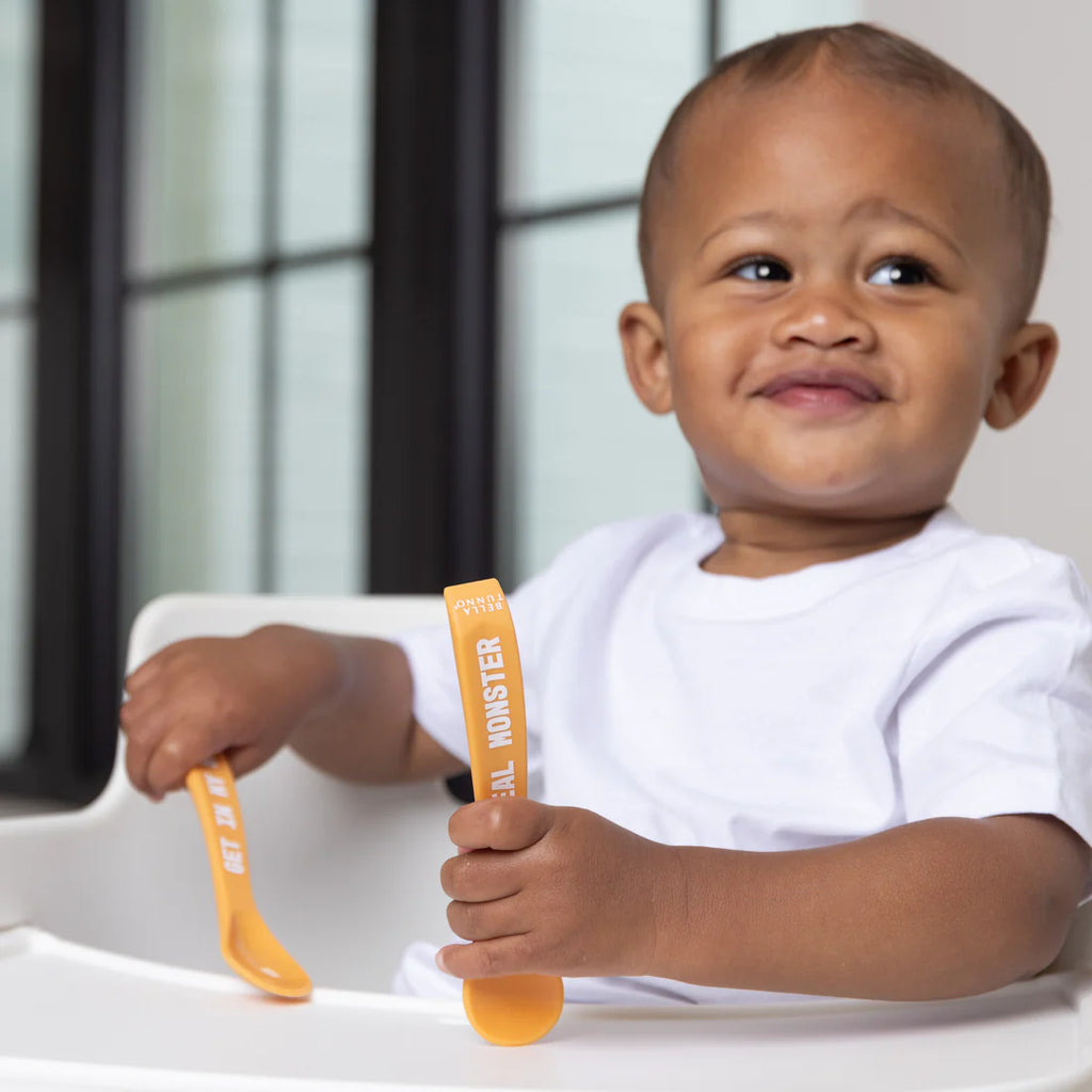 Child holding orange utensils with text, sitting in a high chair.
