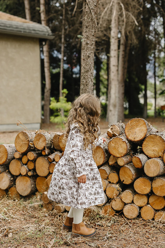 Child in a patterned dress standing next to a stack of logs in a forest setting
