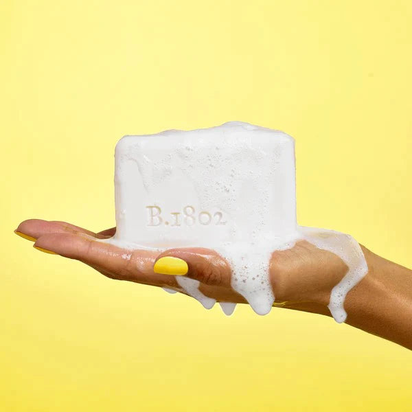 person holding unpackaged bar soap in hand lathered in front of a yellow background
