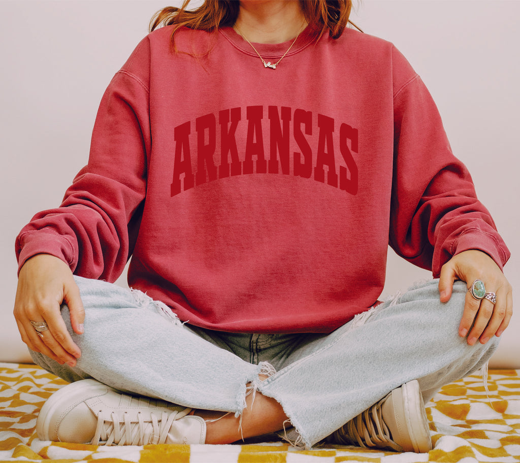 Arkansas crewneck in red with model sitting criss cross with a white background