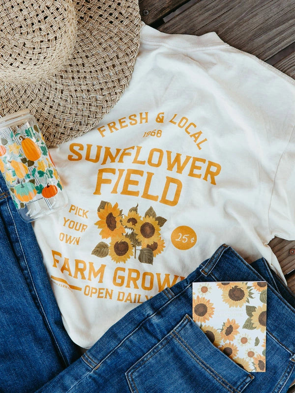White t-shirt with sunflower design and text on a wooden surface with denim jeans and straw hat.