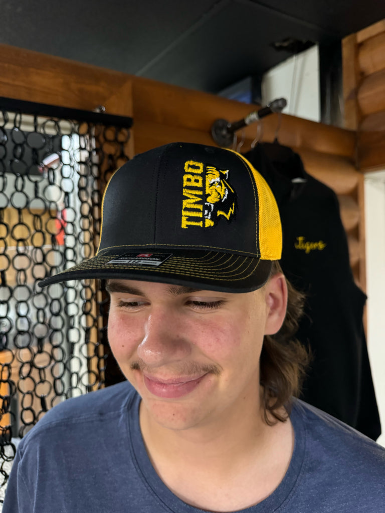 Person wearing a black and yellow cap with 'TIMBO' branding on a patterned floor.