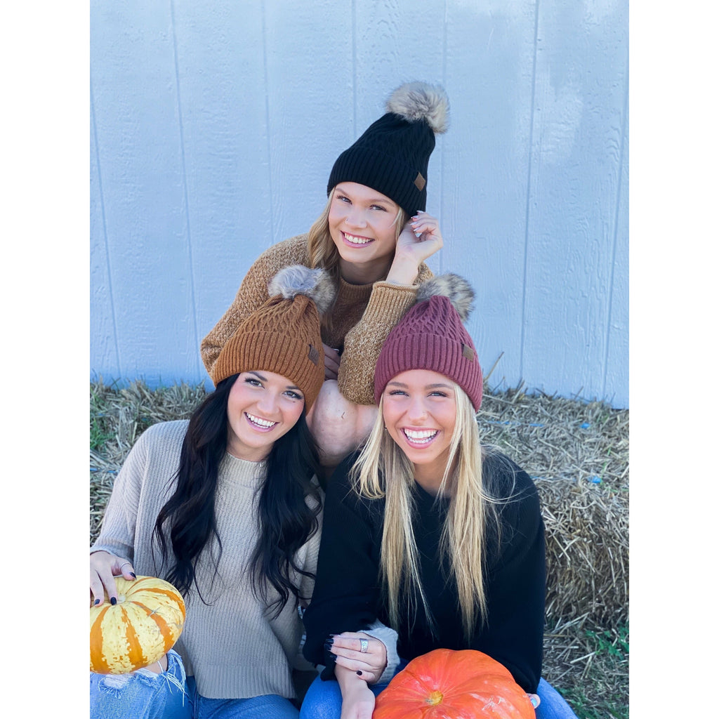 girls wearing solid honey comb fur pom cc beanie in front of white fence and square bail hay
