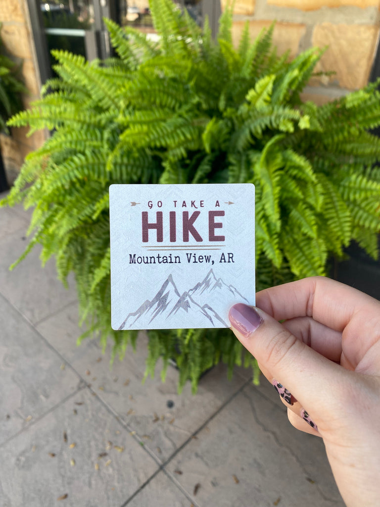 take a hike Mountain View Arkansas magnet being held in front of a green fern
