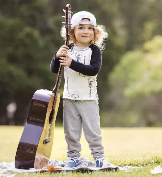 burt's bees babies guitar pant set gray pants and a white top with black sleeves with gray guitars on the shirt worn by a child holding a real guitar in a park with grass and trees